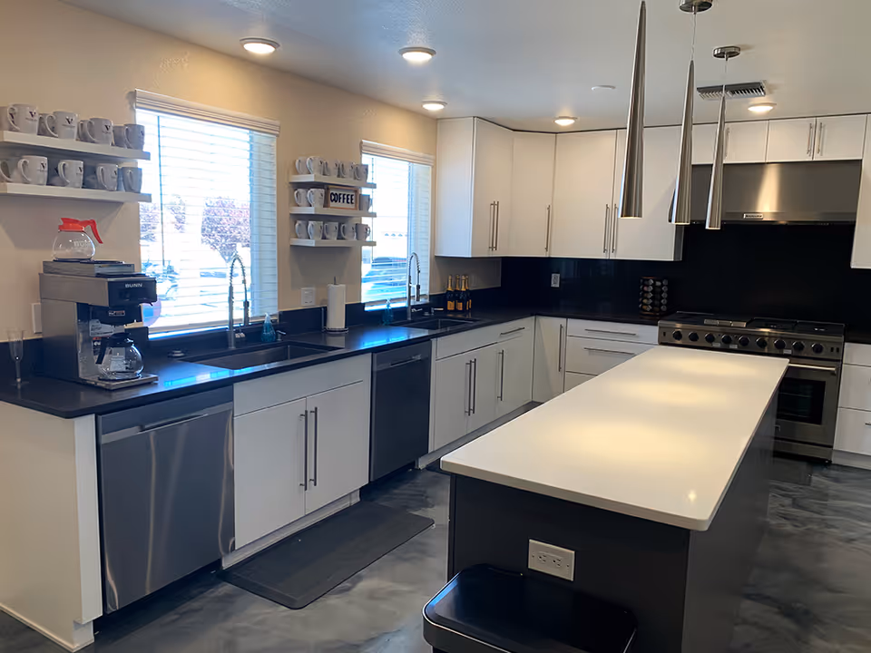 Modern kitchen with white cabinets, black countertops, and stainless steel appliances including a dishwasher and stove. There is a large white island in the center with three pendant lights hanging above it. Two windows with blinds let in natural light, and shelves with coffee mugs and a coffee maker are visible on the left wall.