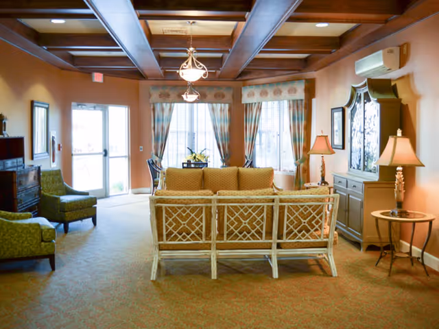 A cozy living room area with a beige sofa facing away from the camera, two green armchairs, a wooden cabinet with lamps on either side, and large windows with patterned curtains letting in natural light. The ceiling features wooden beams and two hanging light fixtures.