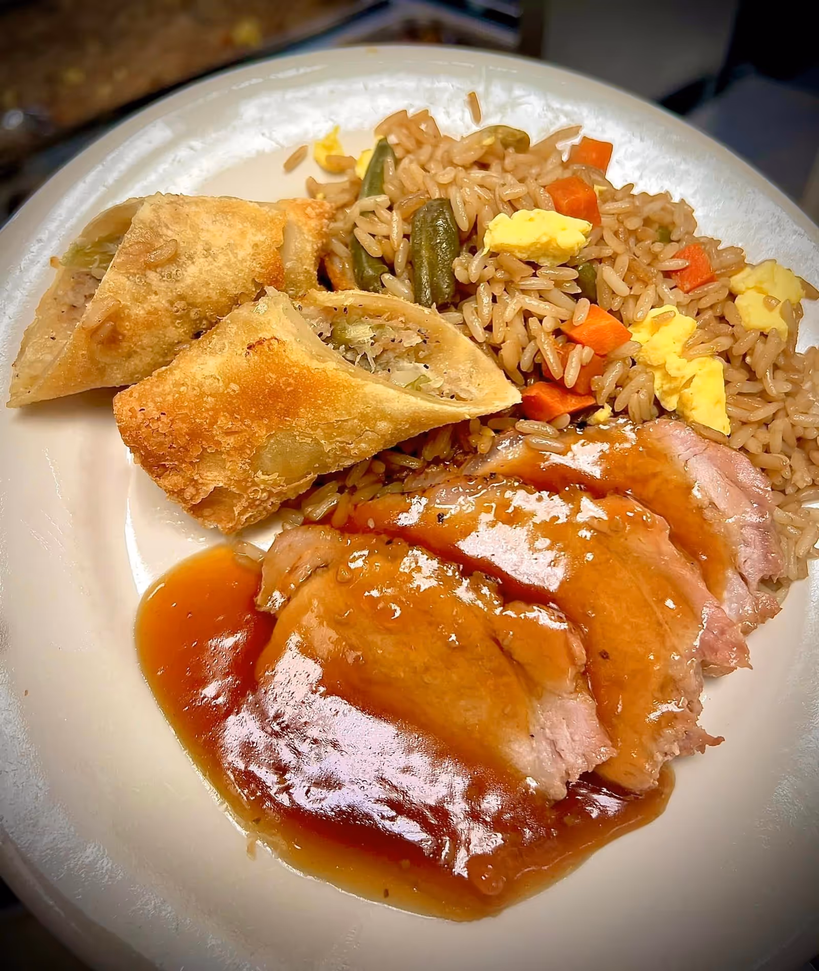 A plate of food with three slices of meat covered in brown gravy, fried rice with vegetables and scrambled eggs, and two fried egg rolls cut in half.