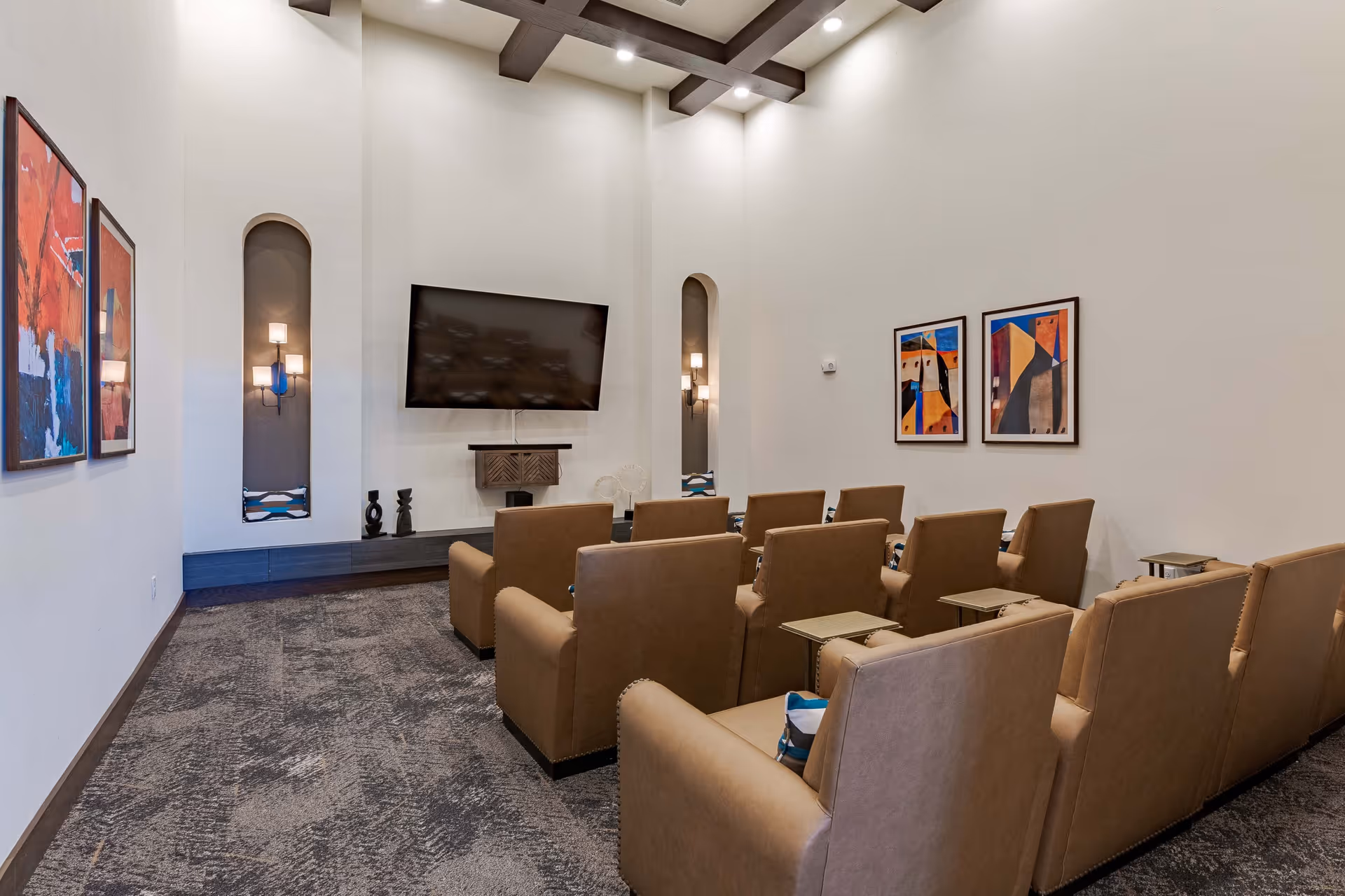 A small theater-style room with two rows of tan leather armchairs facing a large flat-screen TV mounted on a white wall. The room has high ceilings with exposed wooden beams, modern wall sconces in recessed niches, abstract colorful paintings on the walls, and a patterned carpeted floor.