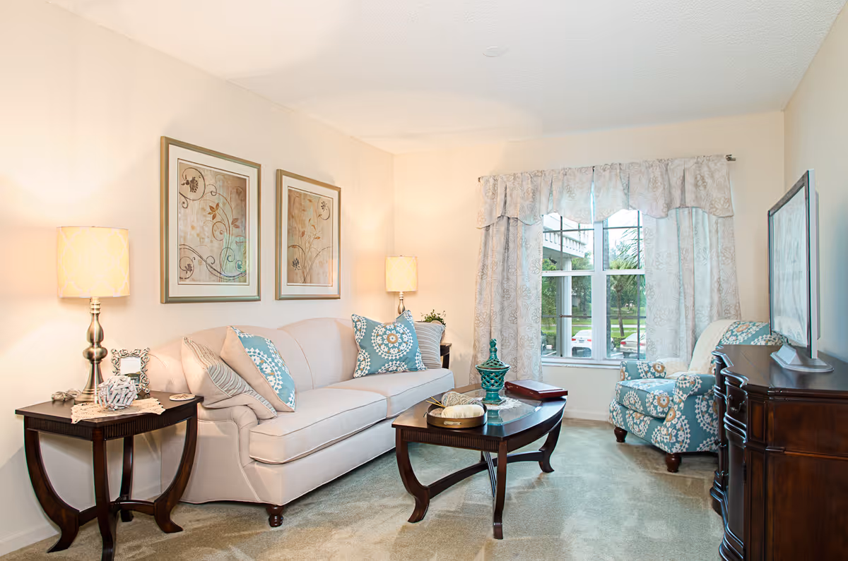 A cozy living room with a beige sofa adorned with patterned blue and beige pillows, two framed floral artworks on the wall above the sofa, a wooden coffee table with decorative items, a matching wooden side table with a lamp and decor, a patterned armchair near a window with light-colored curtains, and a wooden TV stand with a flat-screen TV.