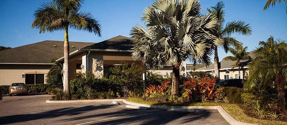 Entrance to a senior living facility with palm trees, shrubs, and parked cars.