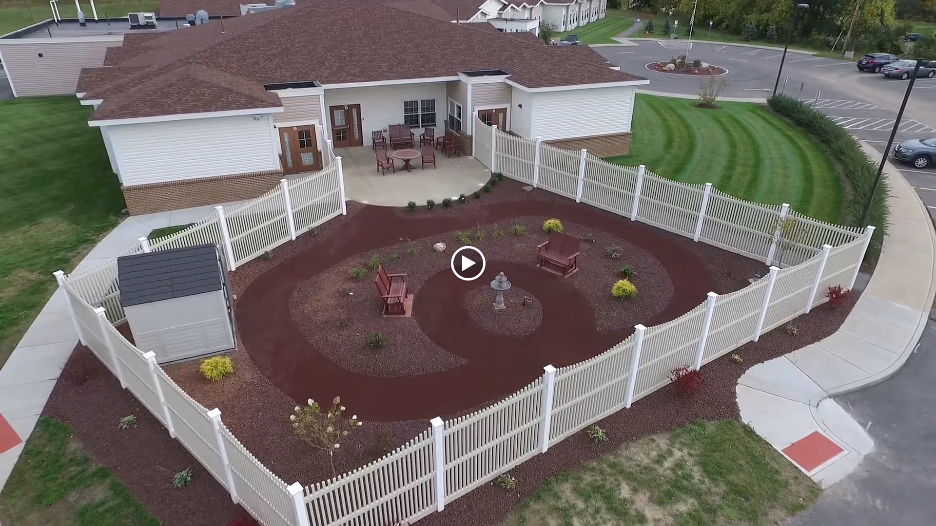 Aerial view of an outdoor fenced garden area at St. Francis Commons Assisted Living Residence. The garden features a circular pathway with two wooden benches and a small birdbath or fountain in the center. Surrounding the garden are small plants and shrubs. Adjacent to the garden is a patio area with several chairs and tables, connected to a building with white siding and a brown roof. The surrounding area includes a parking lot and well-maintained grassy lawns.
