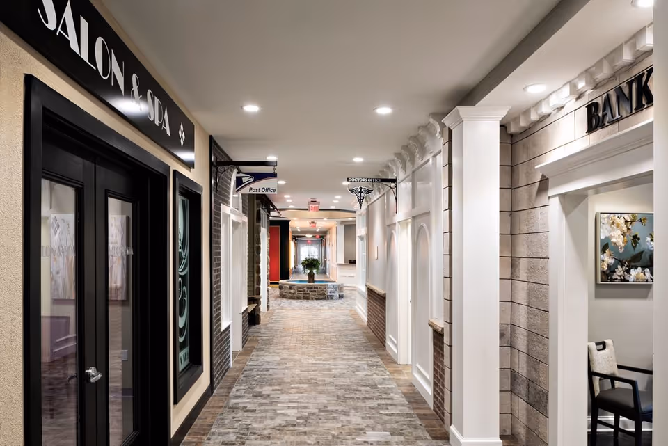 Indoor hallway in a senior living facility designed to resemble a small town street with storefronts including a Salon & Spa, Post Office, Doctor's Office, and Bank. The hallway has a brick-patterned floor, white walls with decorative molding, and recessed ceiling lights. A stone circular bench with a plant is visible at the end of the hallway.