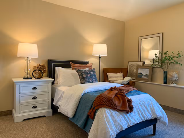 A cozy bedroom with a neatly made bed featuring white and blue bedding, accented with a rust-colored throw blanket and multiple pillows. There is a white nightstand with a lamp and decorative items on the left side of the bed. In the corner, a floor lamp stands next to a brown armchair with a pillow and throw blanket. The room has beige walls and carpeted flooring, with framed pictures and a plant on a ledge by the window.
