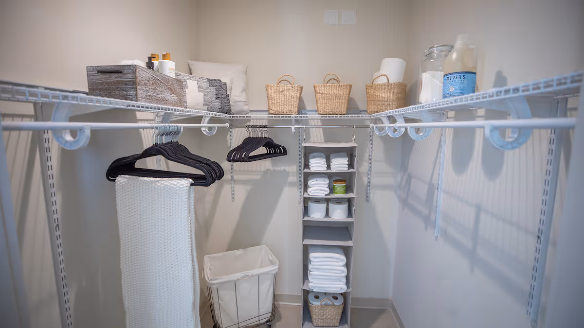 A neatly organized walk-in closet with white wire shelving and hanging rods. The shelves hold woven baskets, folded towels, and various toiletries. Black hangers are hanging on the rods, and a white laundry hamper is placed on the floor.