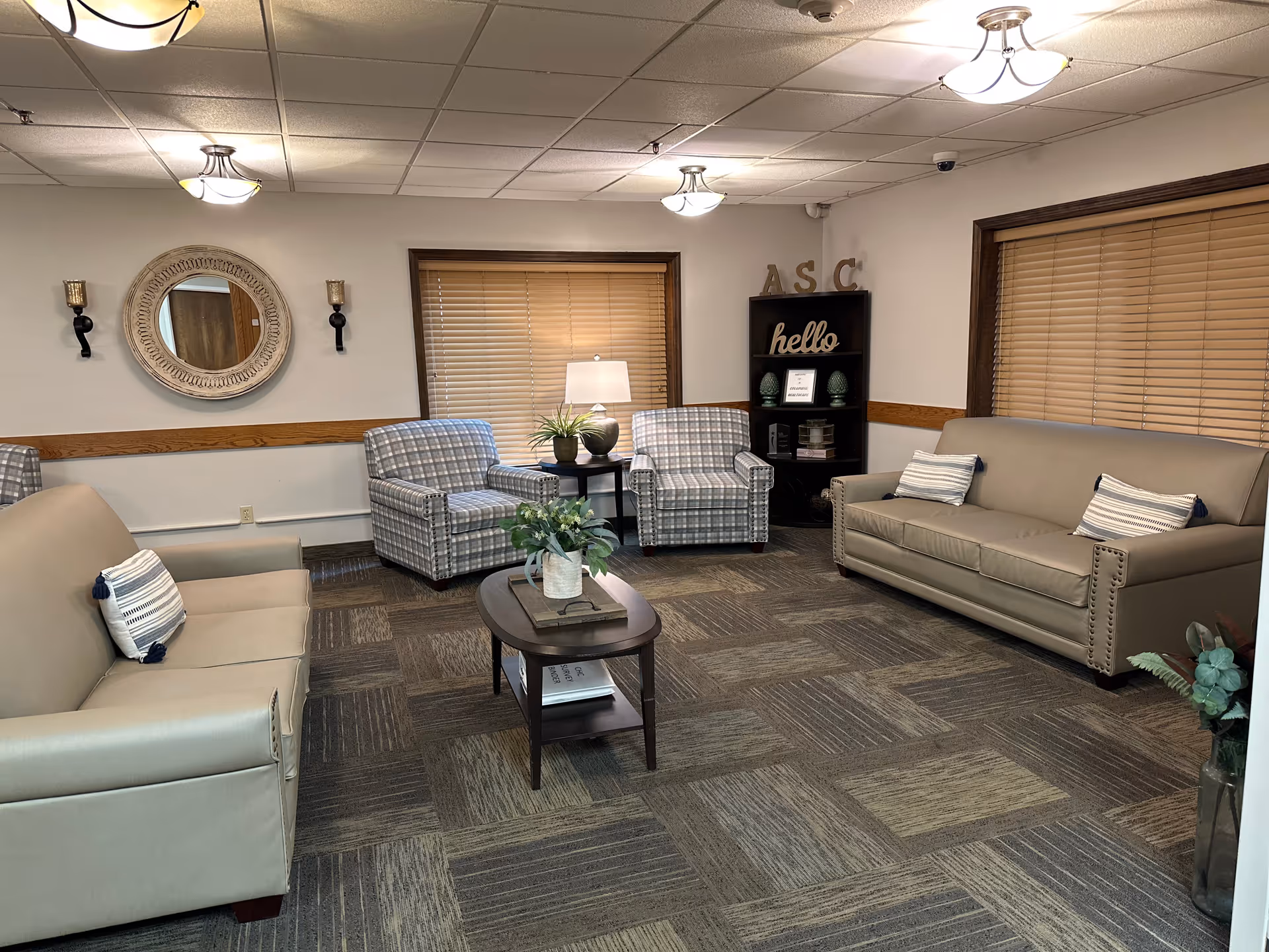 A cozy living room area with two beige sofas, two plaid armchairs, a wooden coffee table with a plant, a round decorative mirror on the wall, and a bookshelf with decorative items and a 'hello' sign. The room has carpeted flooring and windows with wooden blinds.