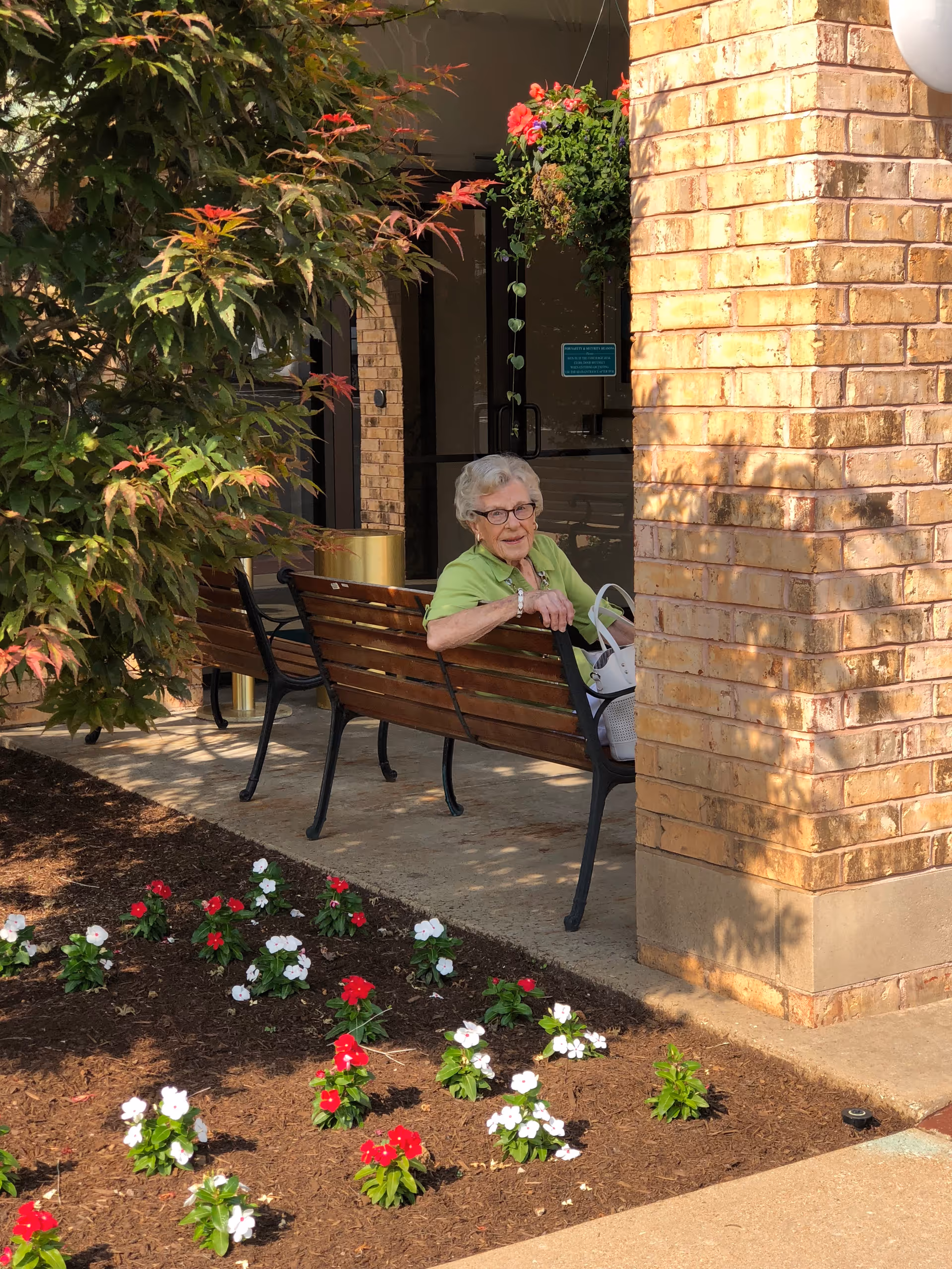 An elderly woman with white hair and glasses sits on a wooden bench outside a brick building. She is wearing a light green shirt and holding a white handbag. There are red and white flowers planted in the garden bed nearby, and a leafy tree partially shades the area.