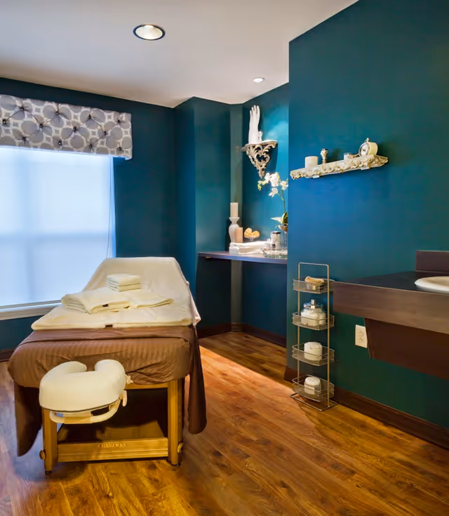 A tranquil treatment room with a massage table covered in linens, teal walls, decorative shelves and a wooden floor.