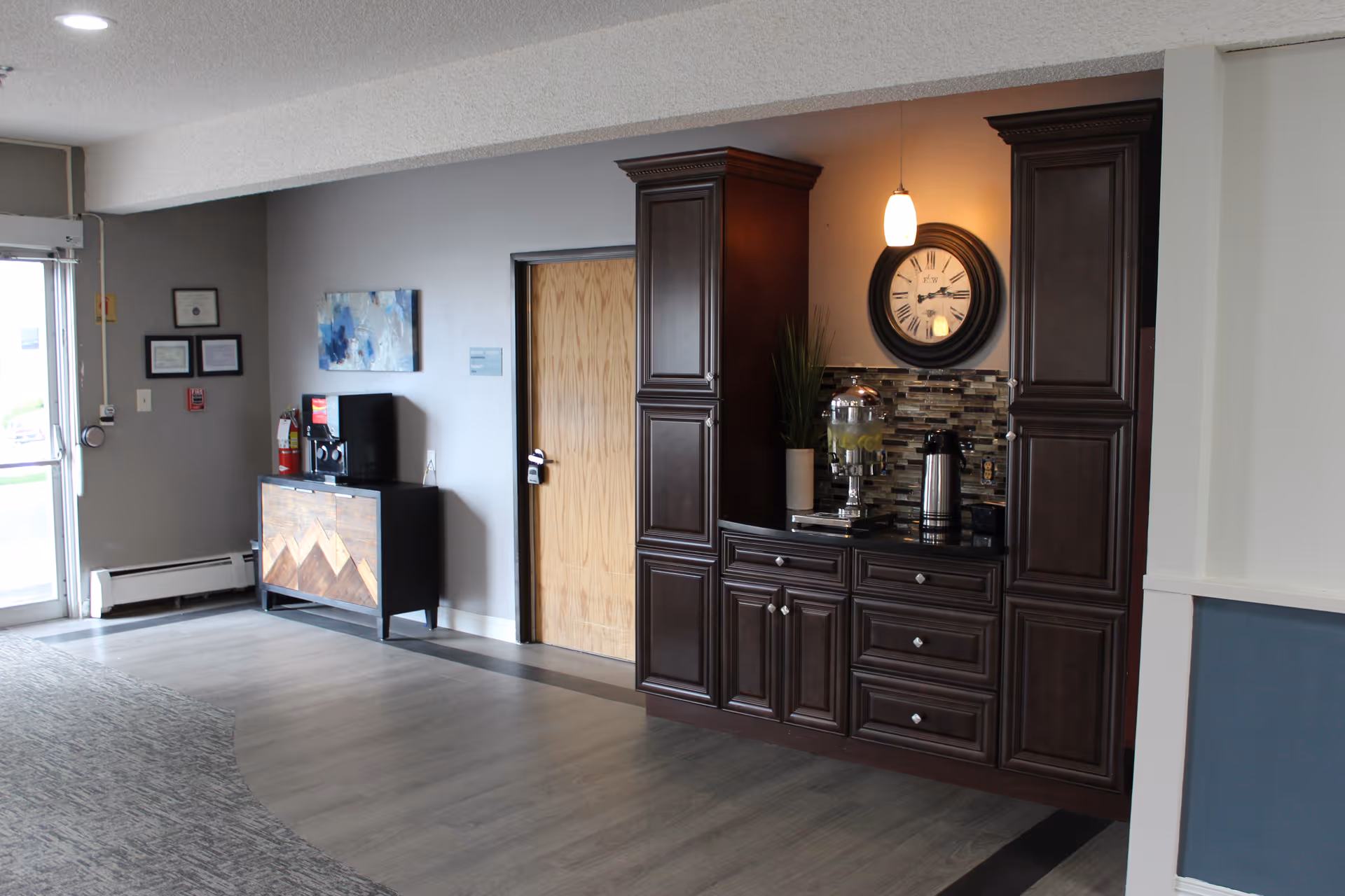 Interior view of a senior living facility hallway with a wooden door, dark wood cabinetry with a clock above, a water dispenser, and a coffee pot. There is a small cabinet with a coffee machine and framed certificates on the gray wall near the entrance door.