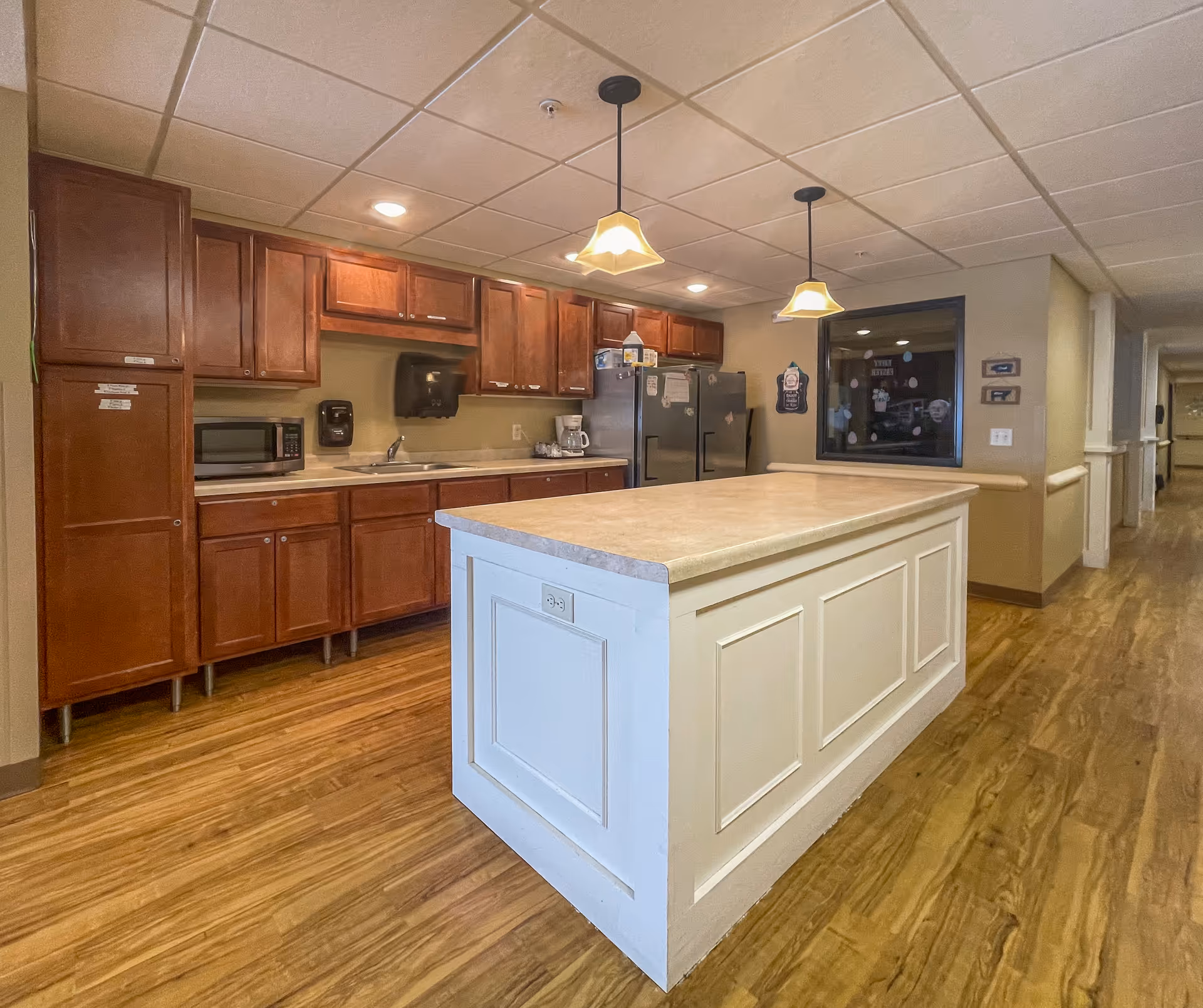 Interior view of a kitchen area in a senior living facility with wooden cabinets, a microwave, a coffee maker, two refrigerators, and a large white kitchen island with a beige countertop. Two pendant lights hang above the island, and there is a hallway visible to the right.