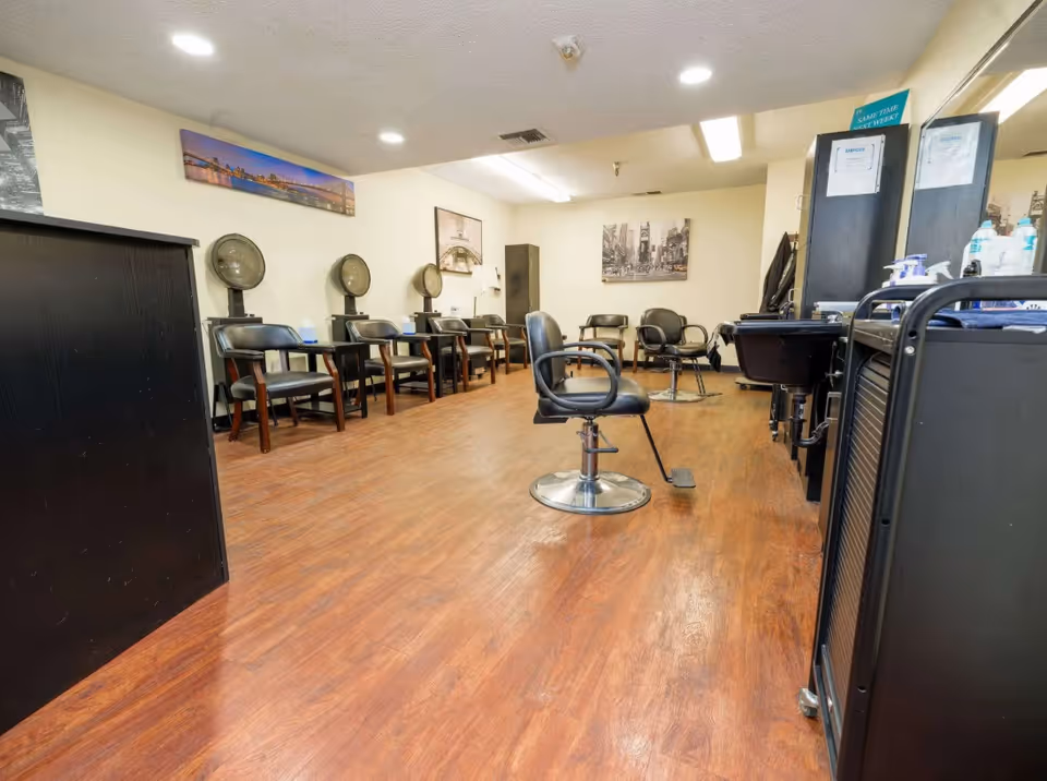 Interior view of a hair salon area in an assisted living facility with multiple black salon chairs, hair dryers mounted on stands, mirrors, and hair washing stations. The room has wood flooring and light-colored walls decorated with framed pictures.