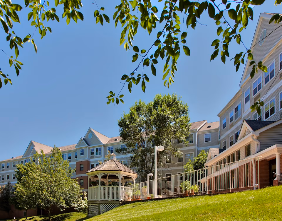 Multi-story senior living building exterior with a wooden gazebo, landscaped lawn, trees, and a clear blue sky.