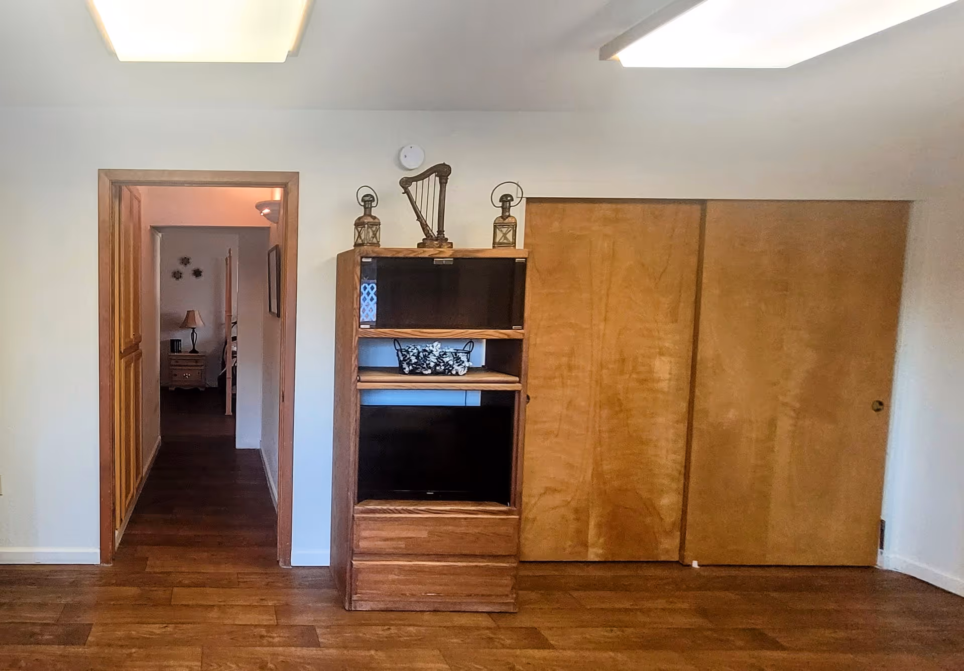 Interior view of a senior living facility room with wooden flooring, a wooden cabinet with decorative items on top, and a hallway leading to another room with a lamp and wall decorations visible.