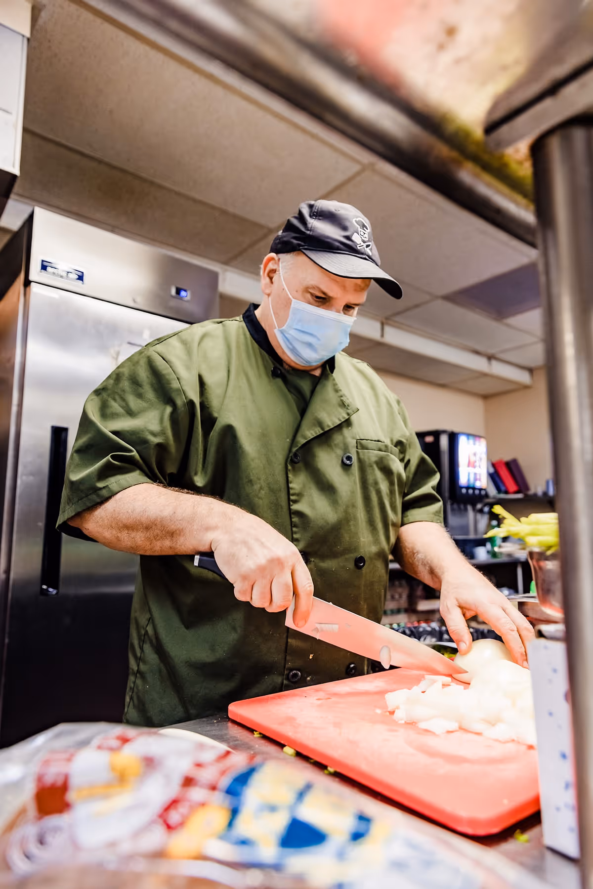 A chef wearing a green uniform, black cap, and blue face mask is chopping onions on a red cutting board in a kitchen with stainless steel appliances and a soda machine in the background.