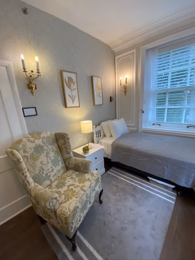 A cozy bedroom with a single bed covered in a gray quilt, two white pillows, and a white headboard. Next to the bed is a white nightstand with a small lamp and a small plant. A patterned armchair with floral upholstery sits on a gray rug with white stripes. The walls are decorated with two framed floral prints and two wall sconces with lit bulbs. A window with white blinds allows natural light into the room.