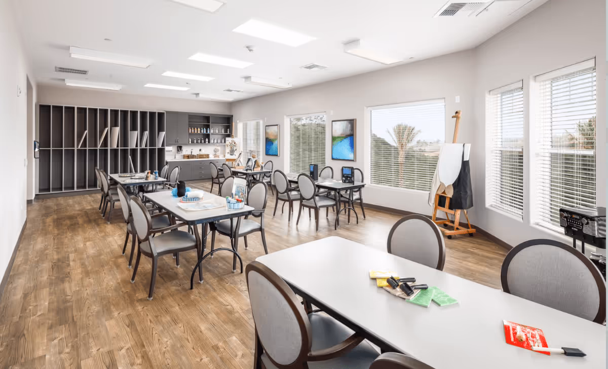 A bright and spacious activity room in a senior living facility with multiple tables and chairs arranged for group activities. The room features large windows with blinds, wooden flooring, and shelves with art supplies. There is an easel with an apron hanging on it near the windows, and various art materials are placed on the tables.