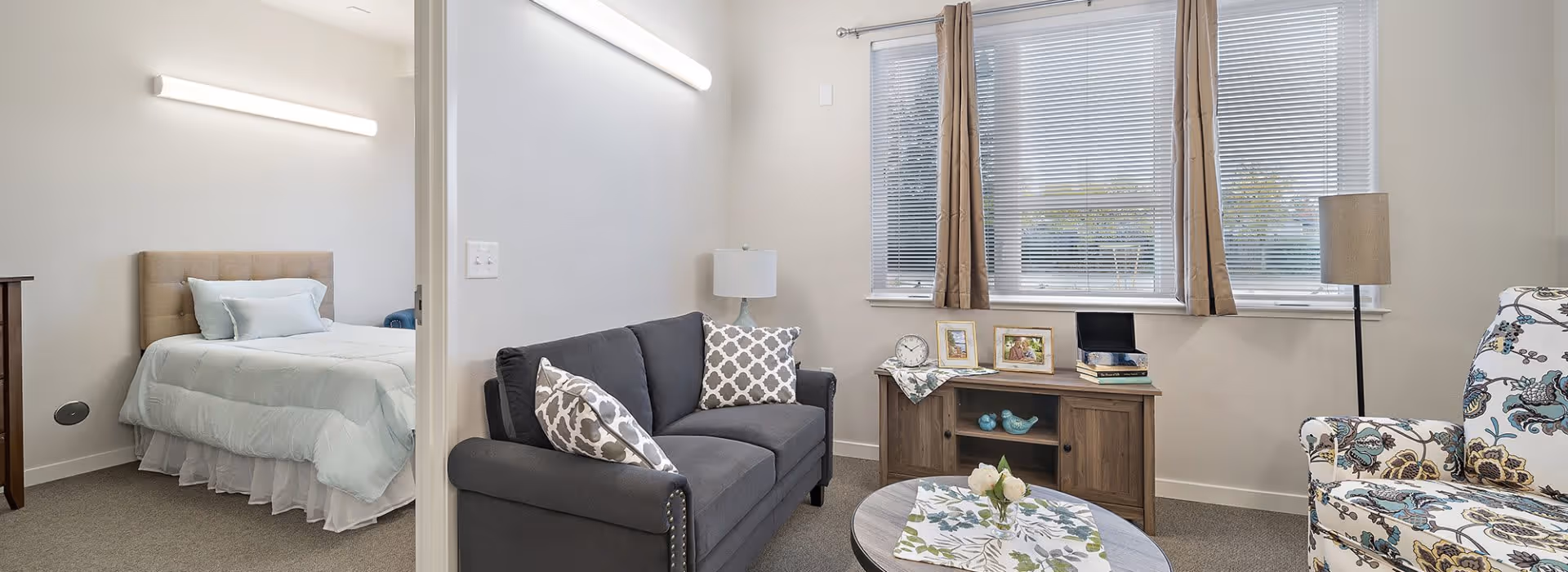 Bright living room with a gray sofa, patterned armchair, coffee table and a bedroom visible through an open doorway.