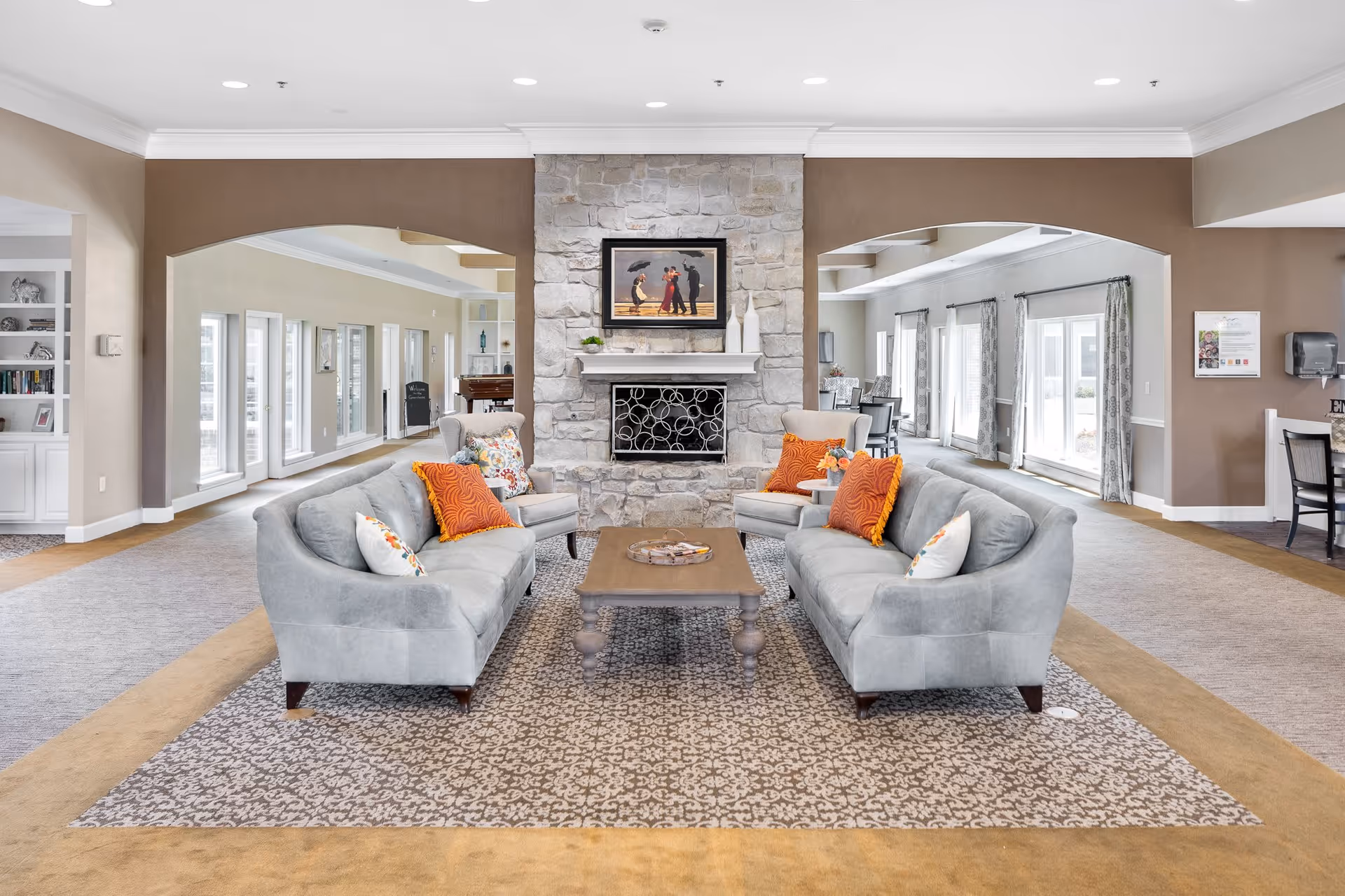A spacious and well-lit living room area with two gray sofas and two armchairs arranged around a wooden coffee table on a patterned rug. The sofas have orange and white decorative pillows. A stone fireplace with a framed painting above it is centered on the far wall. Large windows and archways lead to other parts of the facility, with neutral-colored walls and carpeted floors.