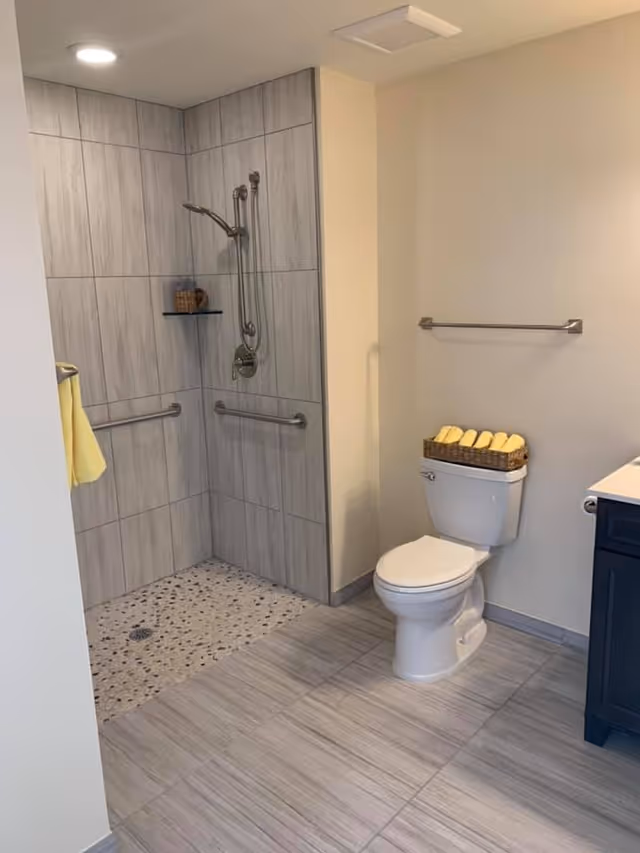 A modern bathroom with a walk-in shower featuring gray tiled walls and pebble stone flooring. The shower has a handheld showerhead and two grab bars. Next to the shower is a white toilet with a basket of rolled yellow towels on top. The floor is tiled in light gray, and there is a towel rack on the wall above the toilet.
