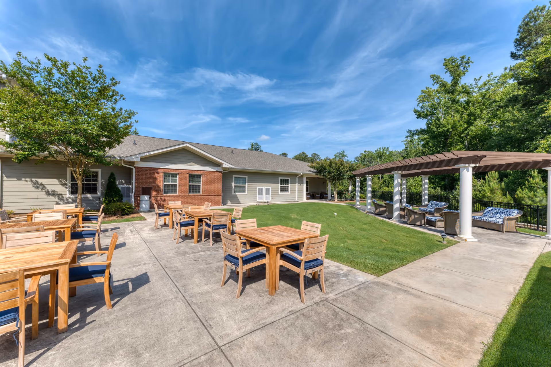 Outdoor patio area at Camellia Walk of Evans with wooden tables and chairs on a concrete surface, a grassy lawn, and a pergola with cushioned seating under a blue sky with some clouds.