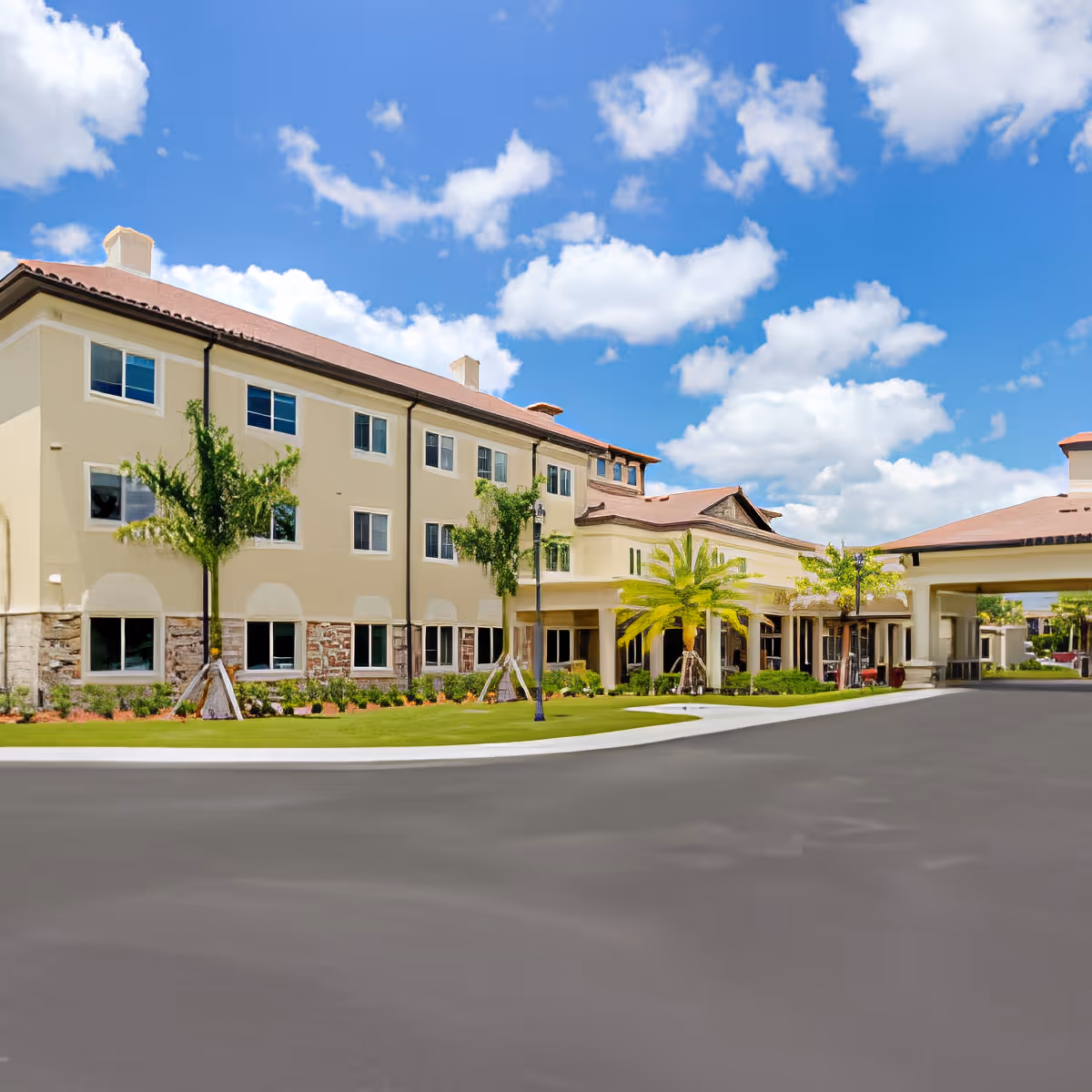 Front exterior of a multi-story senior living building with a porte-cochère, landscaped lawn and palm trees under a blue sky.