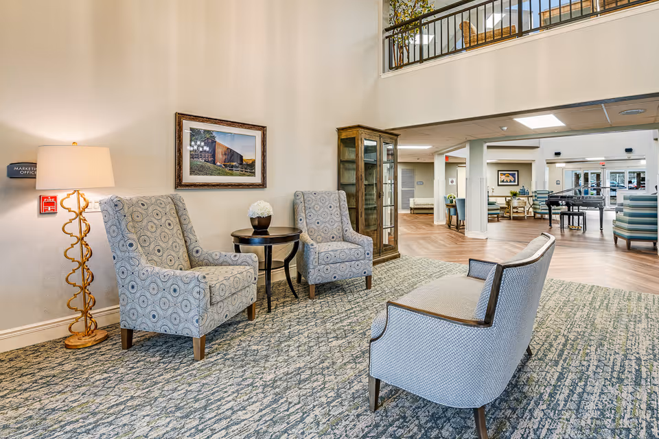 Bright furnished common seating area in a senior living facility with patterned armchairs, a small table and lamp, and an open lounge with a piano in the background.