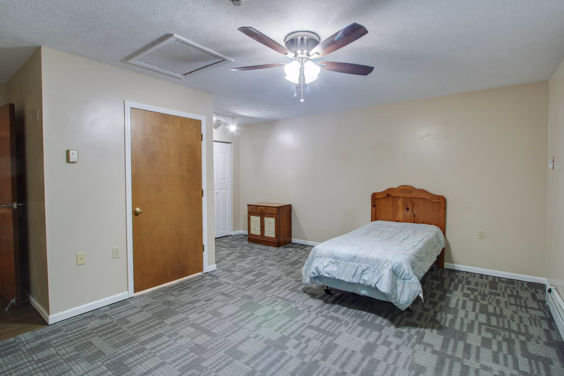 A simple bedroom with a single wooden bed with a light blue patterned comforter, a small wooden cabinet against the wall, beige walls, a ceiling fan with lights, and a patterned gray carpeted floor.