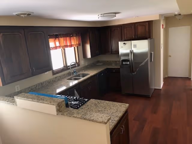A kitchen area with dark wooden cabinets, granite countertops, a stainless steel refrigerator, a gas stove, a double sink under a window with orange curtains, and hardwood flooring.