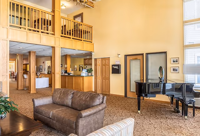 Spacious living area in a senior living facility with a brown leather sofa, a patterned armchair, a black grand piano, and a wooden reception desk. The room features high ceilings, large windows with blinds, wooden railings on an upper level, and beige walls with framed pictures.