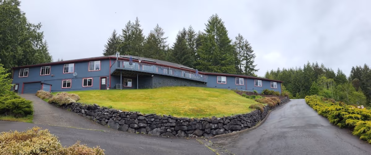 Exterior view of a single-story assisted living facility building with blue-gray siding and red trim, surrounded by green grass, stone retaining walls, and tall evergreen trees under a cloudy sky.