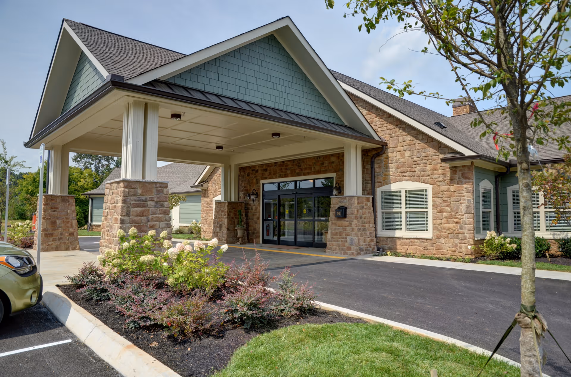 Exterior view of a senior living facility entrance with a covered drop-off area supported by stone pillars, landscaped bushes and flowers, a tree, and a paved driveway leading to glass double doors.