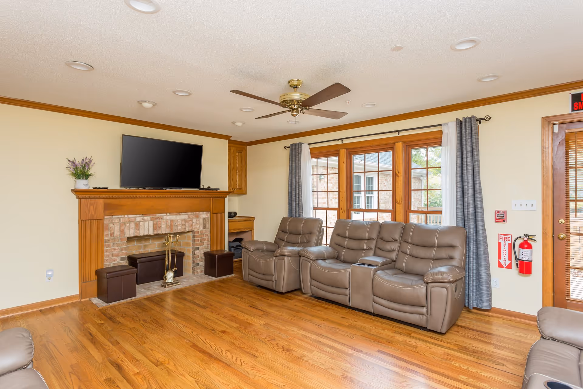 A cozy living room with wooden flooring, a brick fireplace with a wooden mantle, and a flat-screen TV mounted above it. There are three brown leather recliner chairs arranged in front of large windows with gray and white curtains. A ceiling fan with wooden blades is mounted on the ceiling. A fire extinguisher is mounted on the wall near a door with wooden blinds.