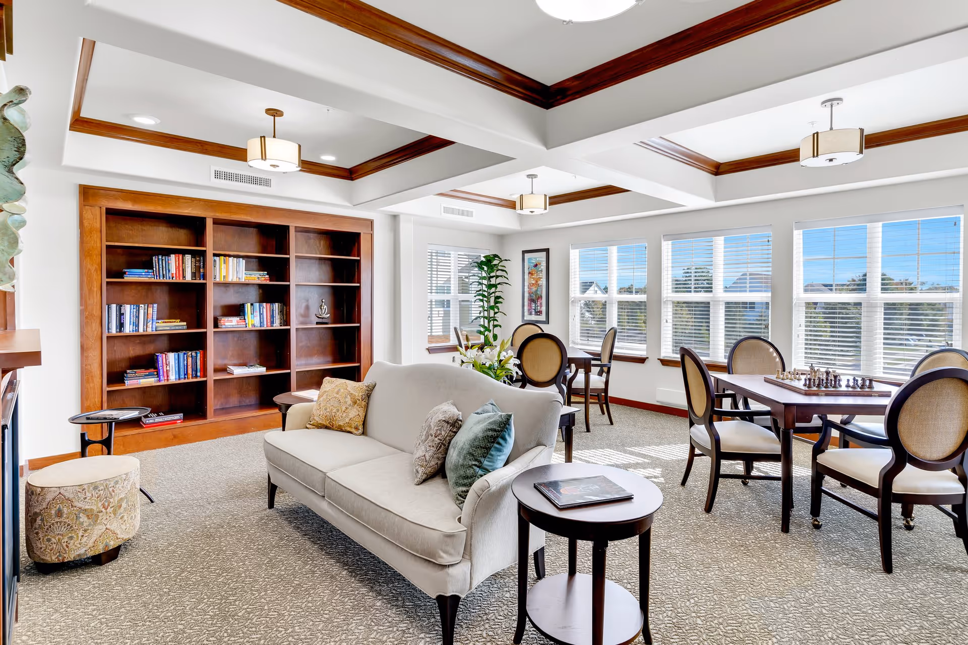 A bright and spacious common area in a senior living facility featuring a beige sofa with decorative pillows, a round side table with a book, a patterned ottoman, and a large wooden bookshelf filled with books. There are multiple tables with chairs near large windows letting in natural light, one table has a chess set on it. The ceiling has recessed lighting and wooden trim accents.