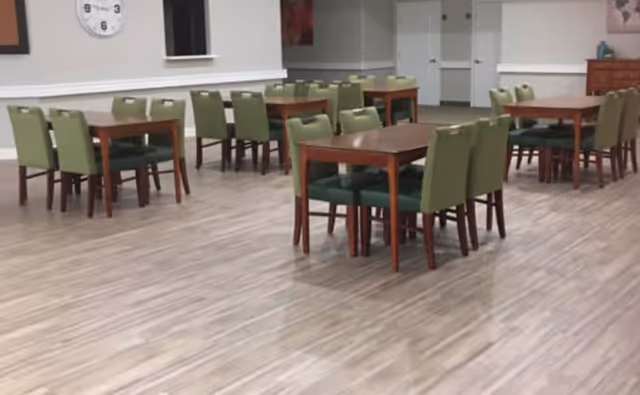 A spacious dining room with several wooden tables and green cushioned chairs arranged neatly on a light wood floor. The walls are light-colored with a clock and some artwork visible, and a doorway is seen in the background.