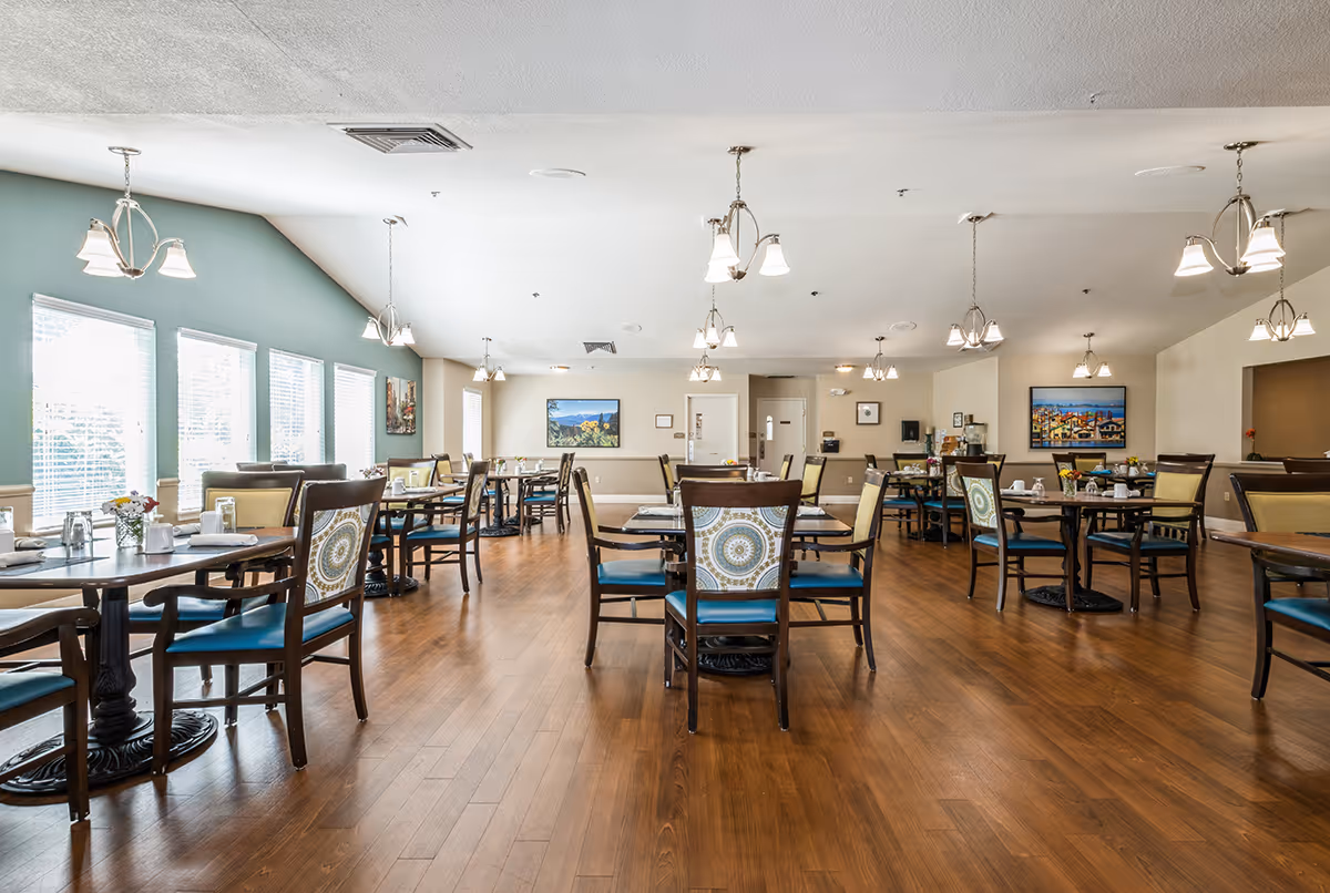 Spacious, well-lit dining room with multiple round tables and chairs, large windows on the left, and pendant lights overhead.
