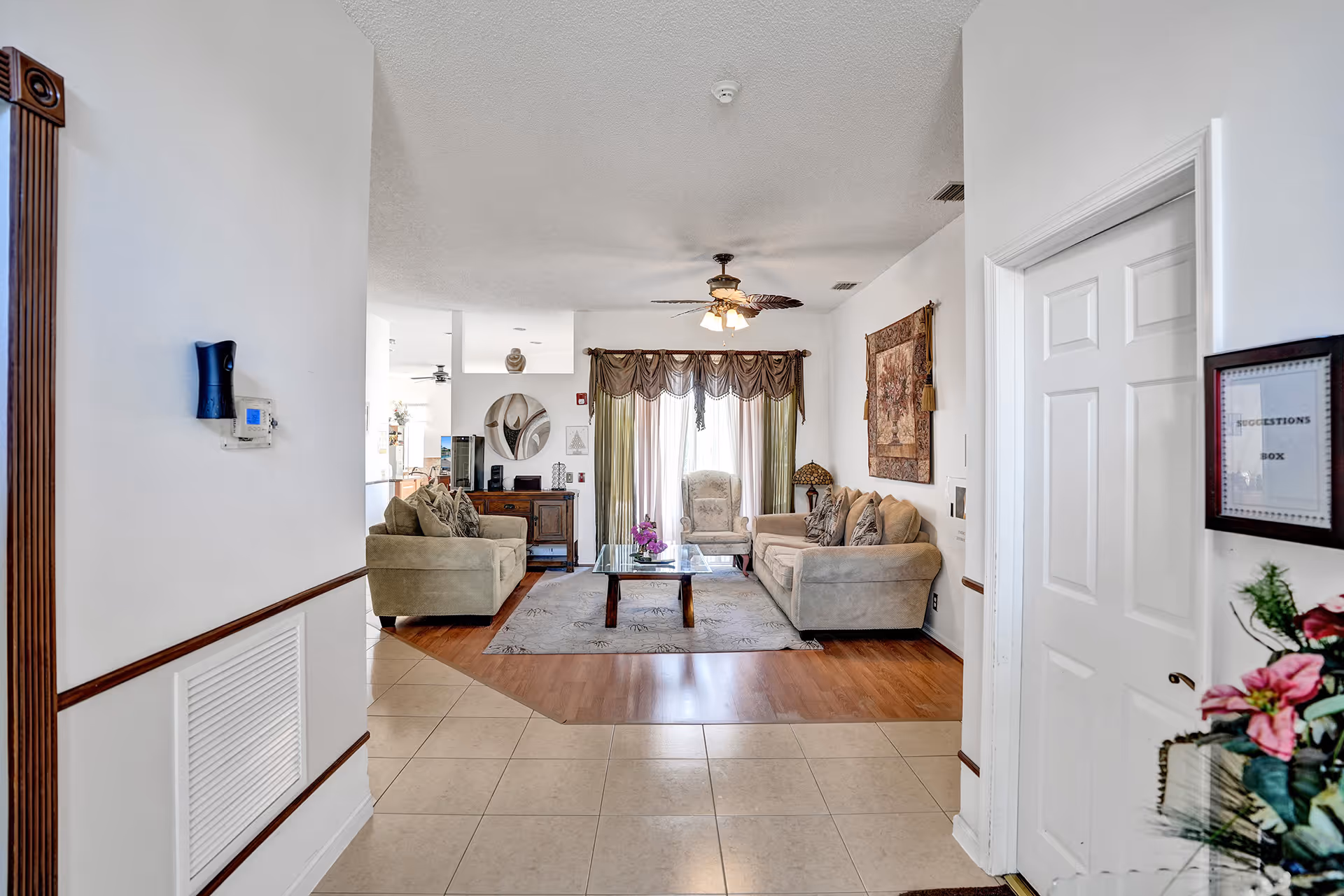 A cozy living room area with two beige sofas, a glass coffee table on a patterned rug, an armchair near a window with curtains, a ceiling fan with lights, a wall tapestry, and a suggestion box mounted on the wall near a white door. The floor transitions from tiled to wood in the living area.