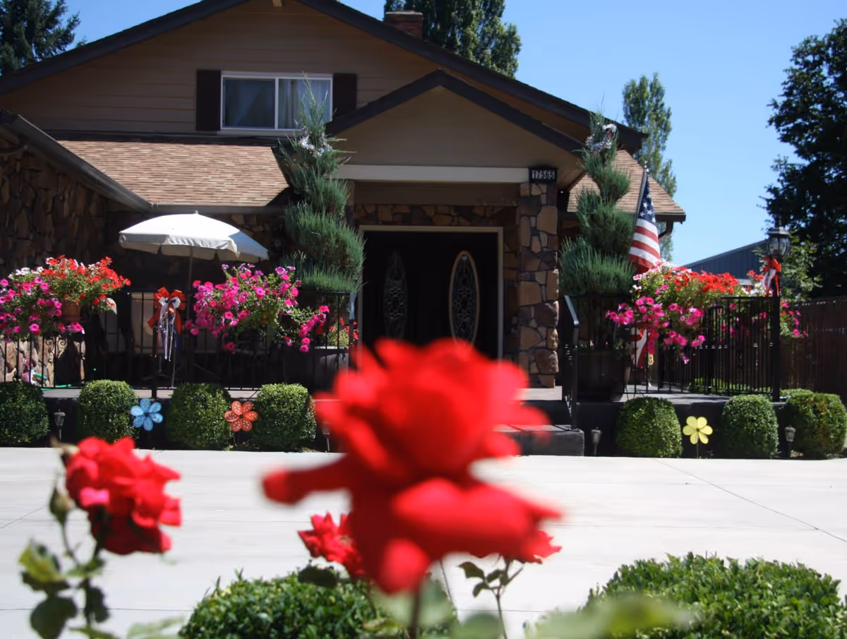 Front entrance of a house with a porch decorated with colorful hanging flowers, potted shrubs, an American flag, and blurred red roses in the foreground.