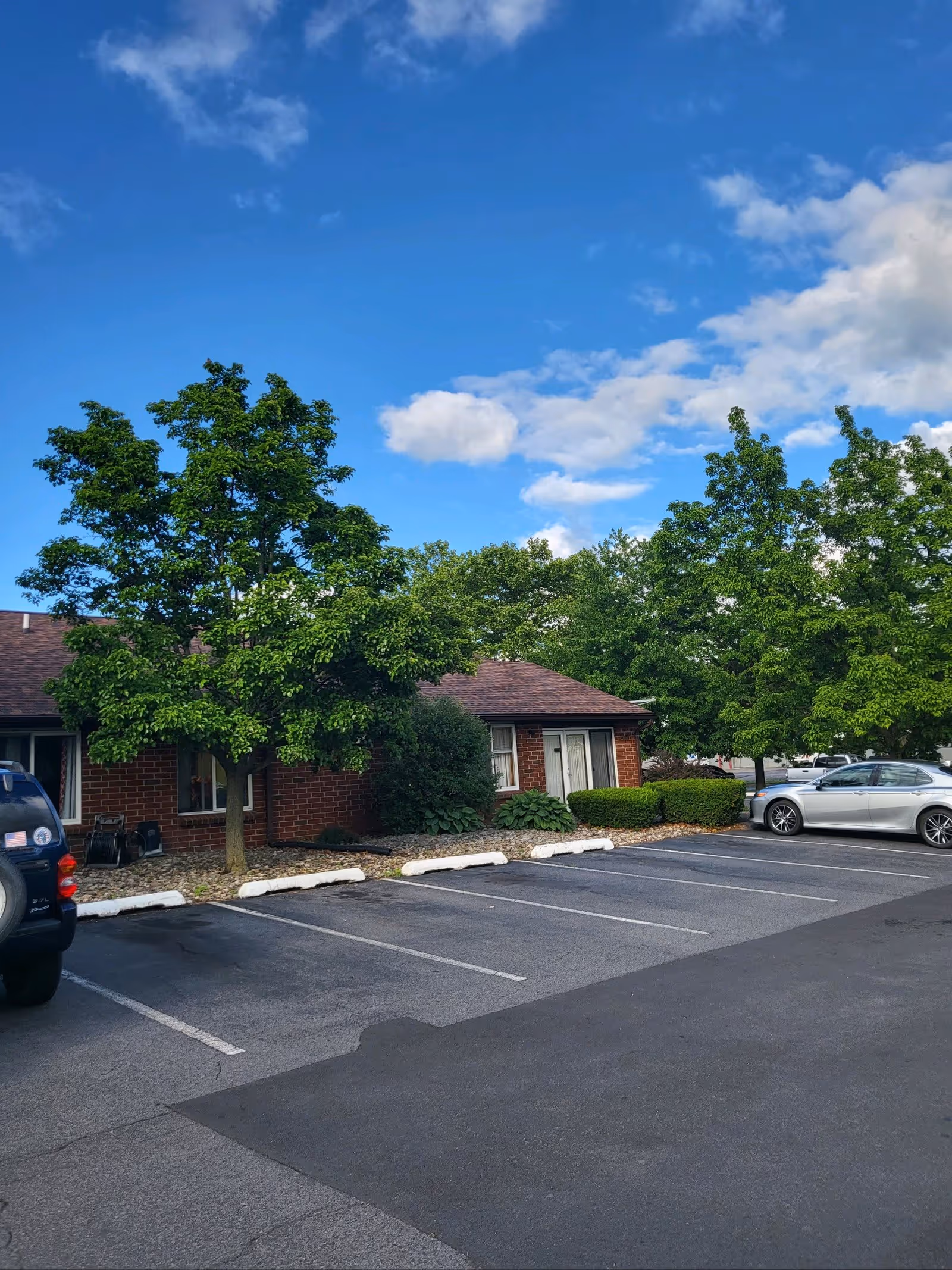 Parking lot with several empty parking spaces and two parked cars in front of a single-story brick building with a brown roof. There are green trees and bushes around the building under a blue sky with some clouds.