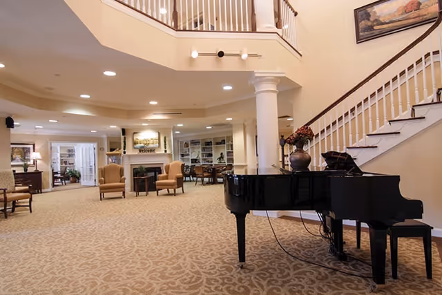 Spacious senior living lobby with a grand piano, seating area, fireplace, staircase, and bookshelves.