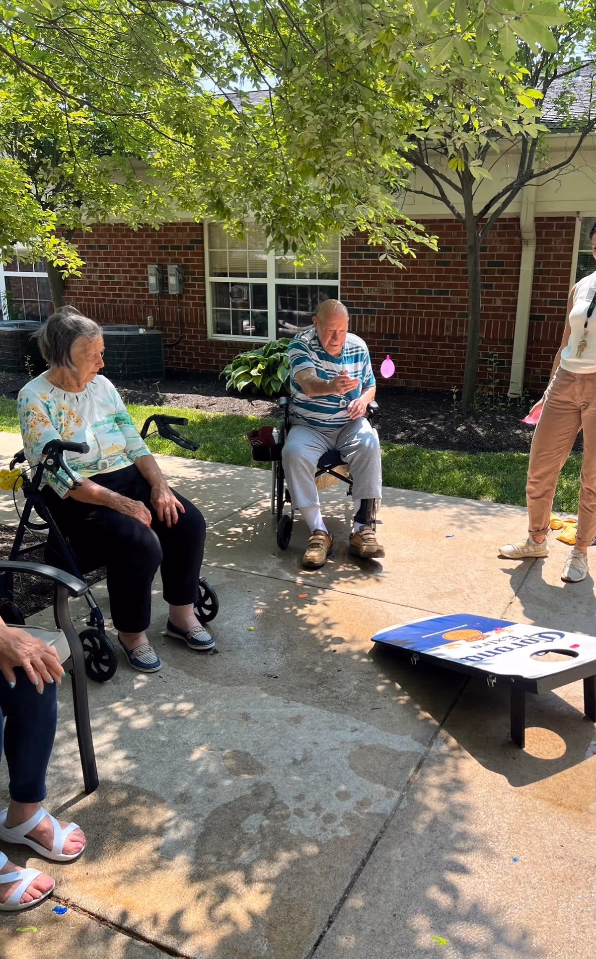 Three elderly individuals sitting outside on a sunny day near a brick building with windows. One man in a wheelchair is playing a game of cornhole, tossing a bean bag towards the board. Two women are seated nearby, one on a walker with a seat and the other on a chair. Trees provide shade over the concrete patio area.
