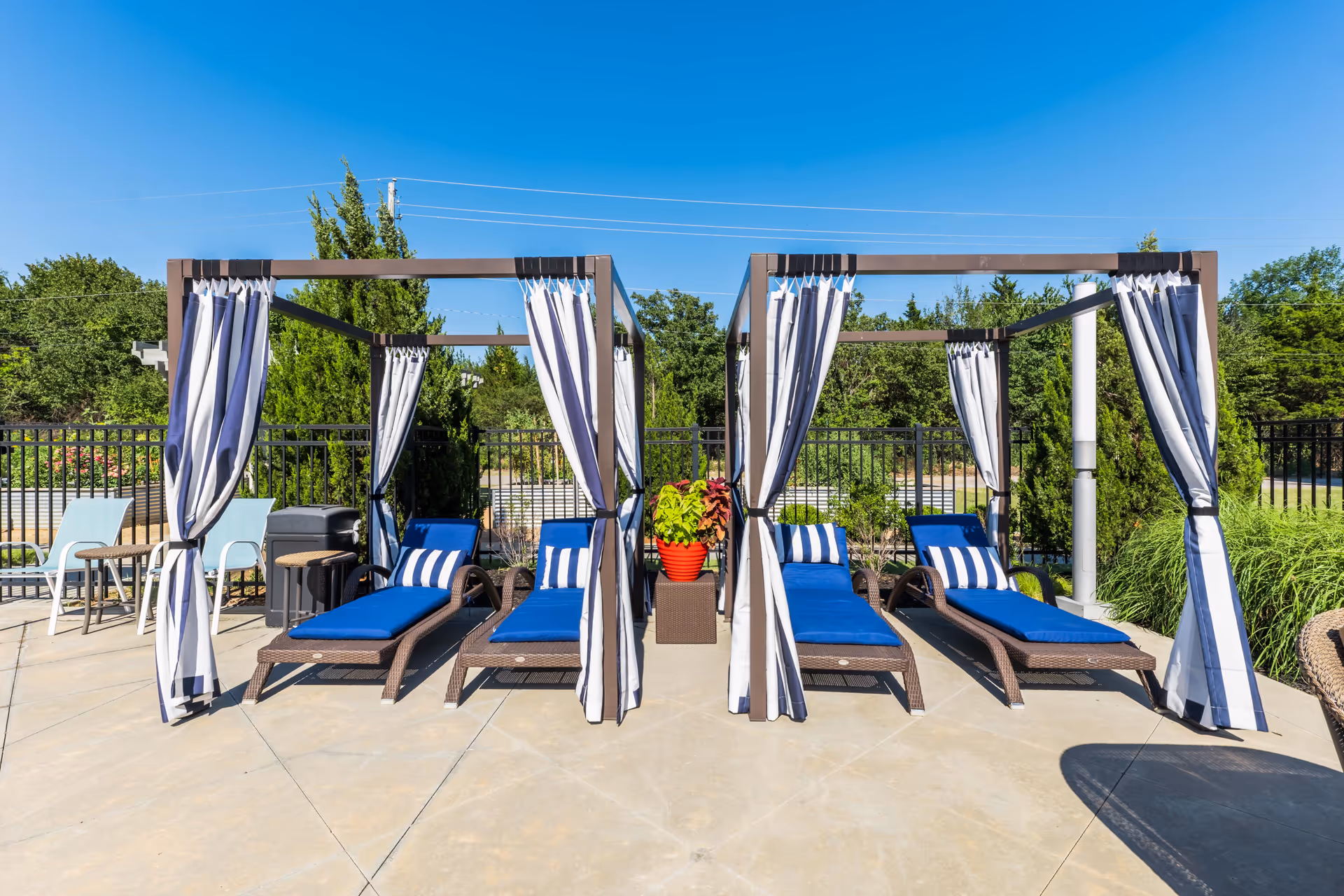 Outdoor patio area with two cabanas featuring blue cushioned lounge chairs and striped curtains, surrounded by greenery and a clear blue sky.
