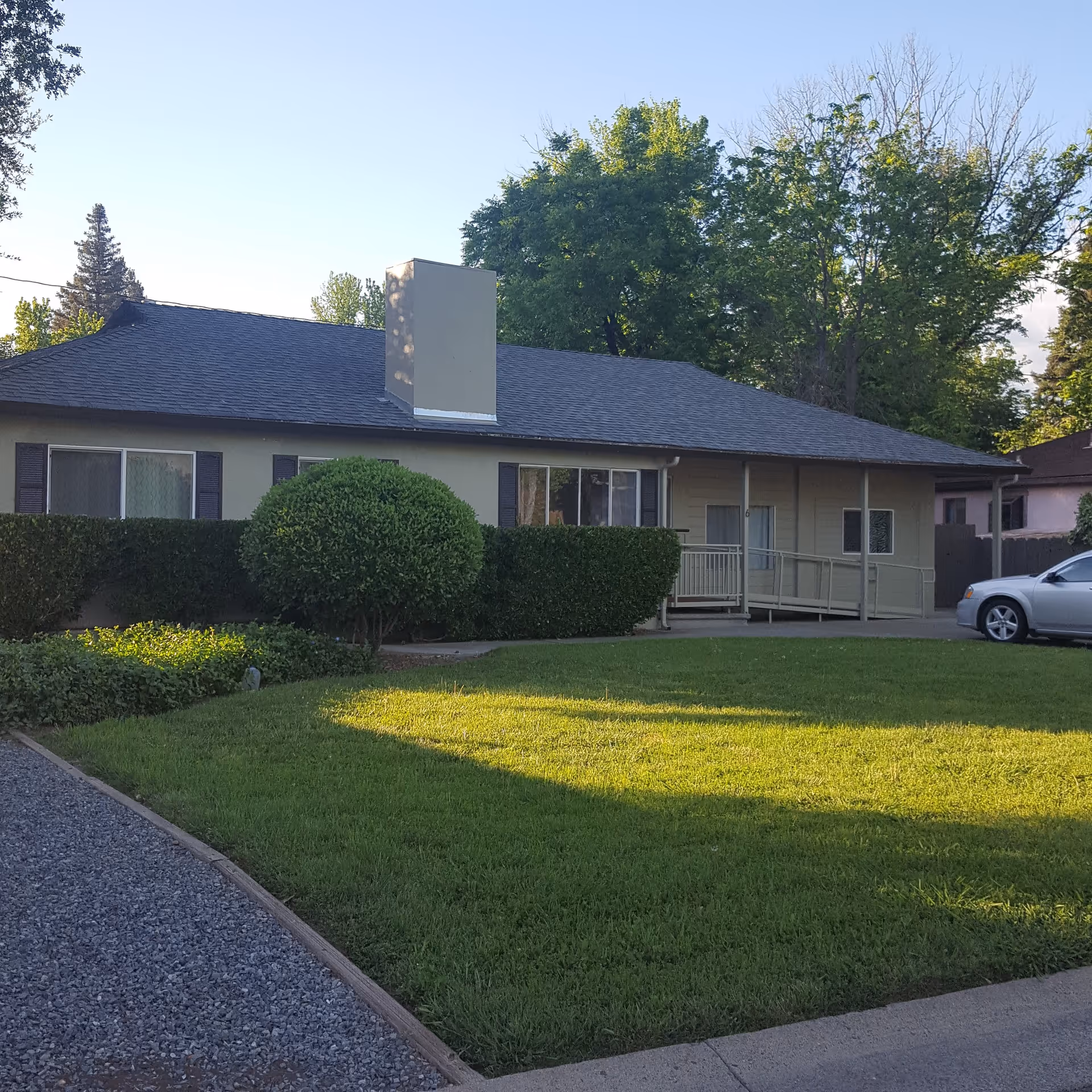 Single-story building with beige exterior walls, dark roof, and a chimney. The front yard has a well-maintained green lawn, trimmed bushes, and trees in the background. A gravel pathway and a concrete driveway with a silver car parked are visible.