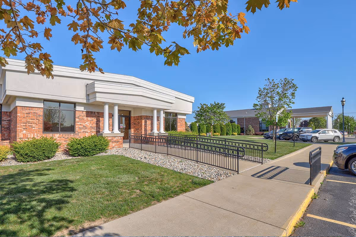 Entrance of a single-story brick nursing and rehab building with a wheelchair ramp, columns, landscaped lawn, and parked cars under a clear blue sky.
