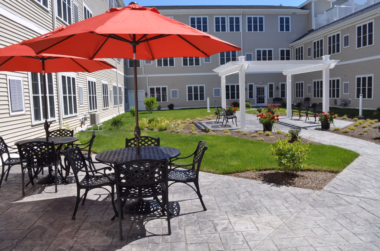 Sunny courtyard with black metal tables and red umbrellas, a white pergola, flower beds, and a surrounding beige multi-story building.