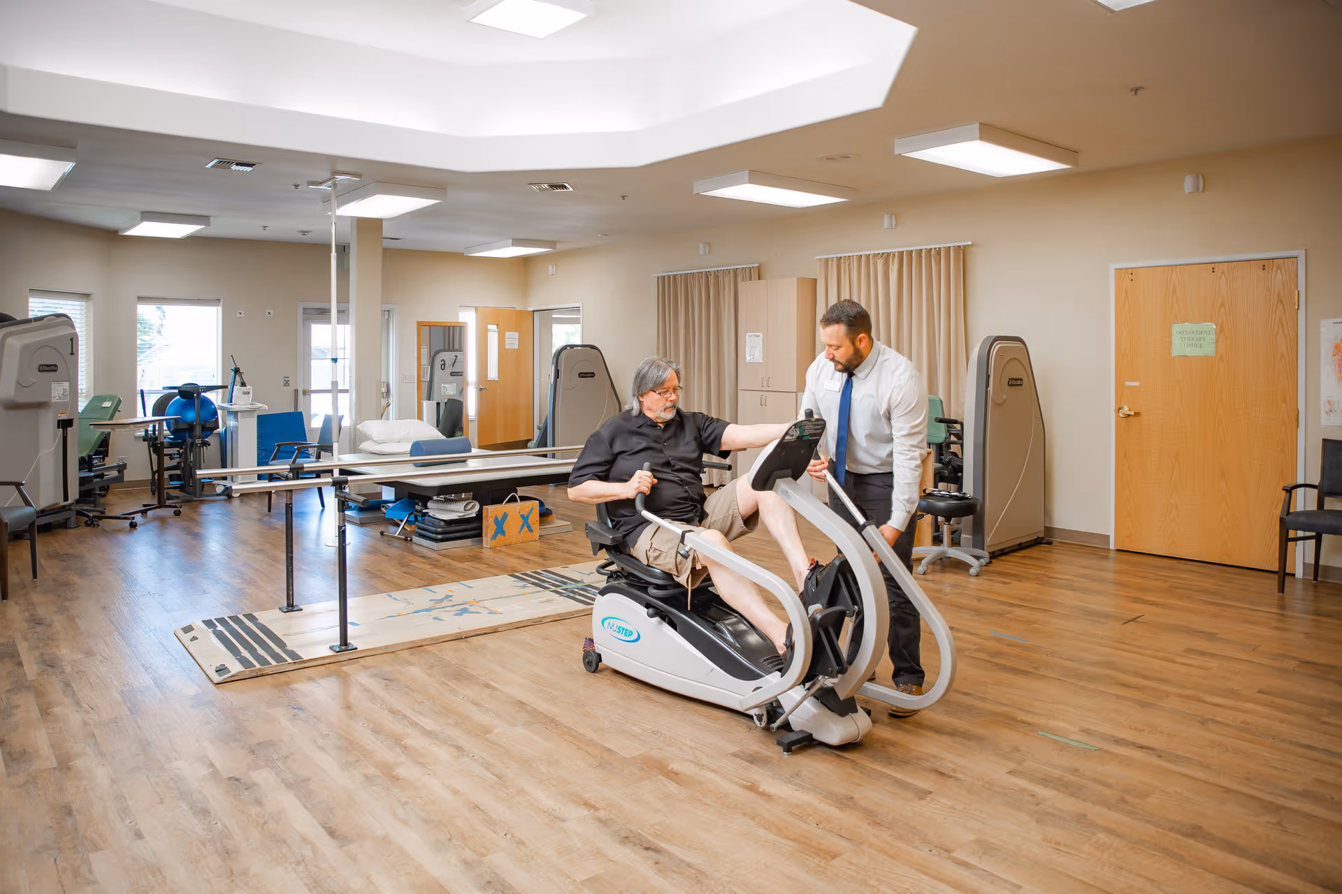 A senior man exercises on a recumbent bike in a spacious rehabilitation or physical therapy room while a male therapist in a white shirt and tie assists him. The room has wooden floors, various exercise and therapy equipment, and large windows letting in natural light.