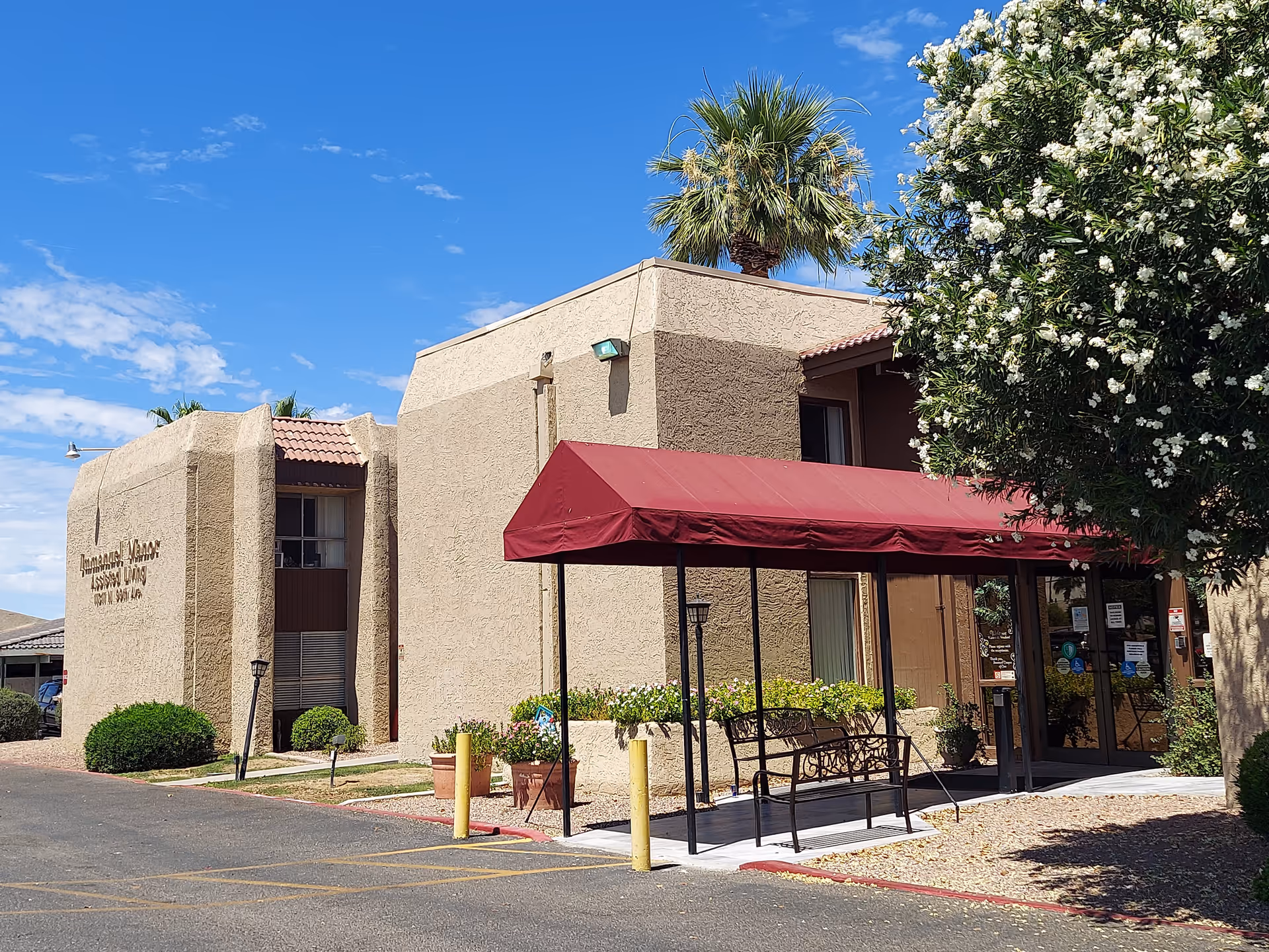 Exterior view of Immanuel Manor Assisted Living building with beige stucco walls, a red canopy over the entrance, a bench underneath the canopy, and a palm tree and flowering bush nearby under a blue sky with scattered clouds.