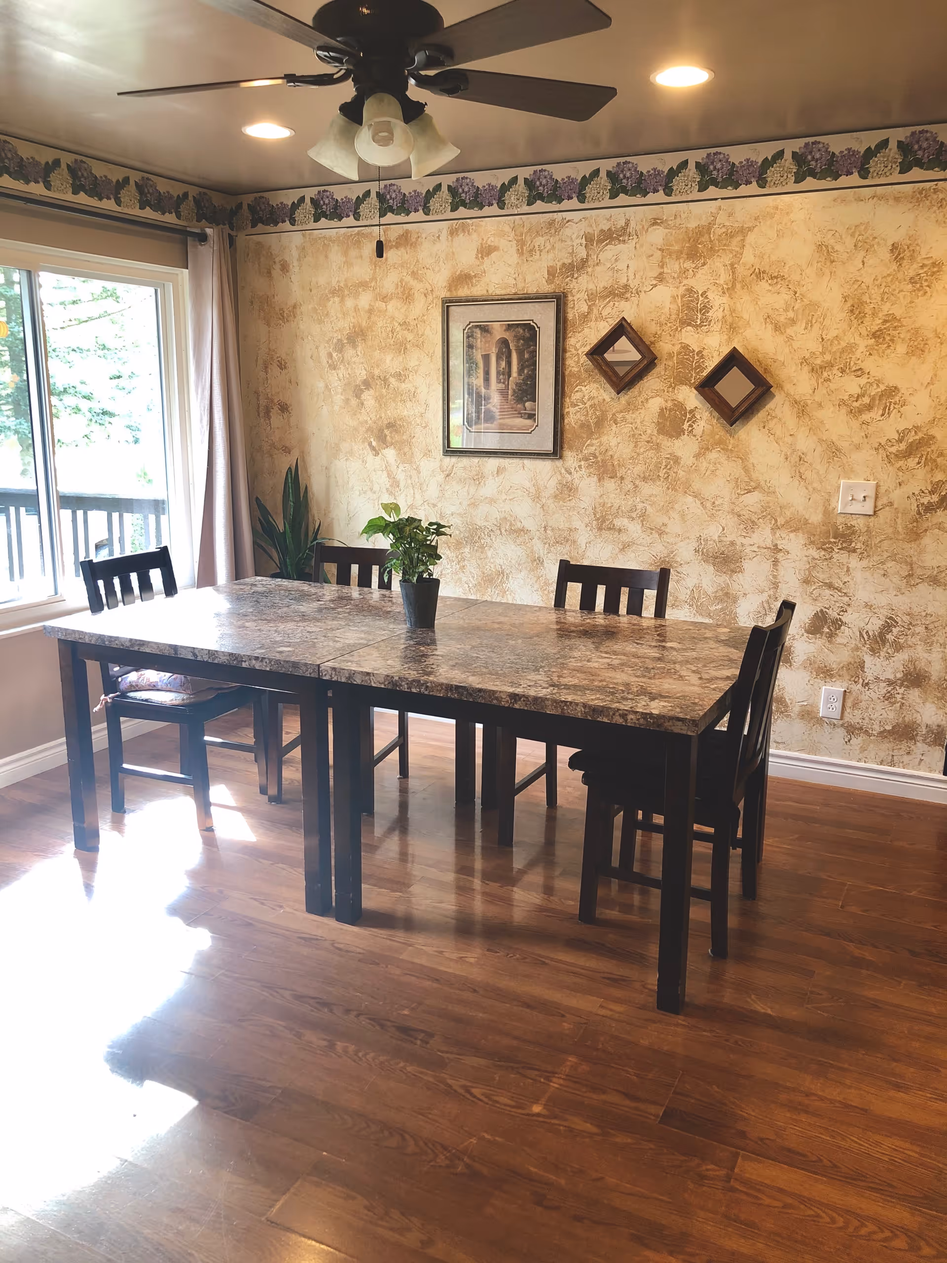 A dining room with a rectangular table and six chairs. The table has a marble-like surface with a small potted plant in the center. The room has wooden flooring, a ceiling fan with lights, and a large window with curtains letting in natural light. The walls have a textured beige finish with a grapevine border near the ceiling and three decorative wall hangings.