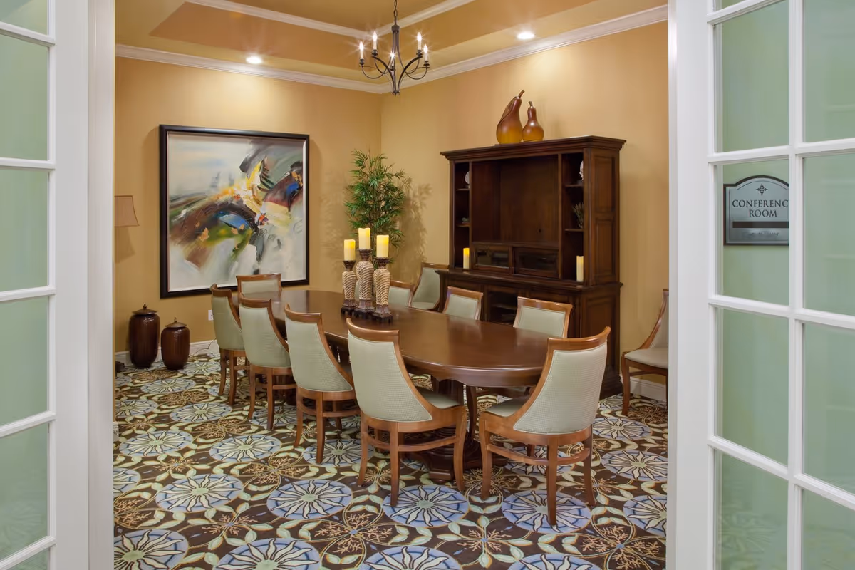 A conference room with a long wooden table surrounded by ten upholstered chairs. The room has a patterned carpet with floral designs, beige walls, and a large abstract painting on one wall. There is a wooden cabinet with decorative items and candles, and a potted plant in the corner. The room is viewed through partially open glass-paneled double doors with a sign that reads 'Conference Room'.