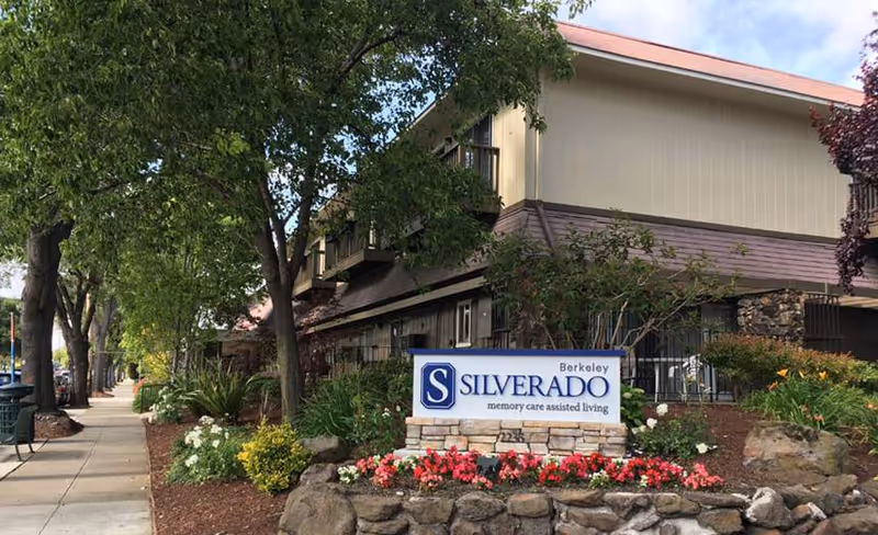 Front exterior of Silverado Berkeley Memory Care Community showing the facility sign in a stone planter, landscaped flowers, trees, and a tree-lined sidewalk.